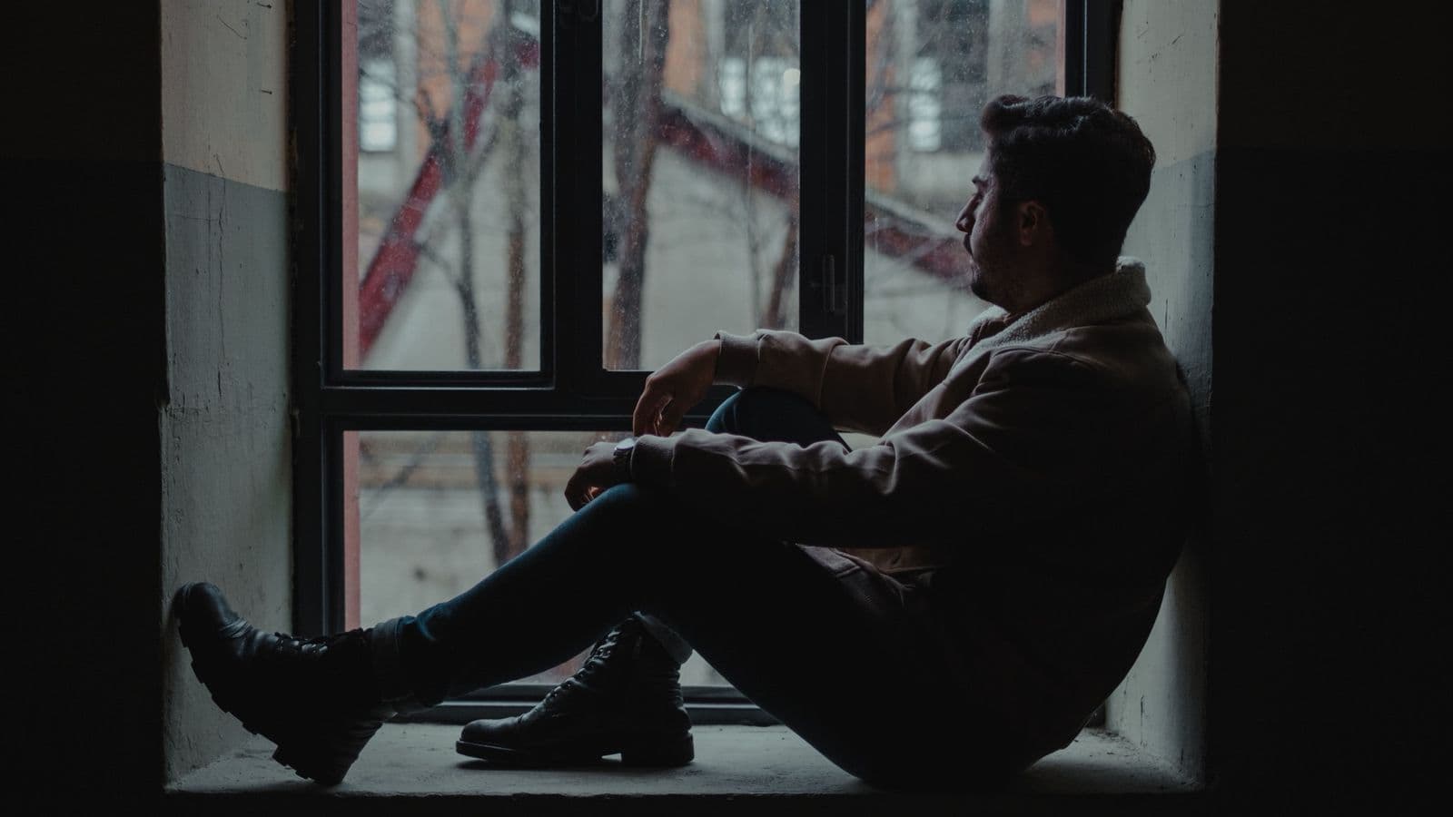 A thoughtful man sits by a rain-streaked window in a dim room