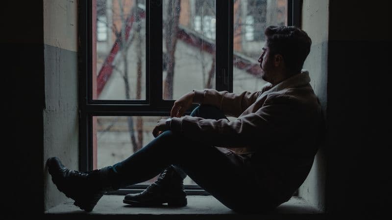 A thoughtful man sits by a rain-streaked window in a dim room