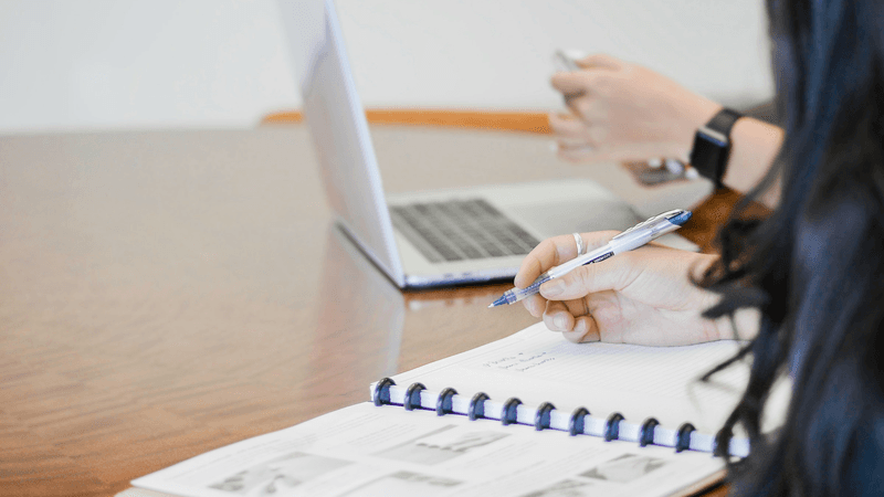 A person writes notes beside a laptop and printed documents