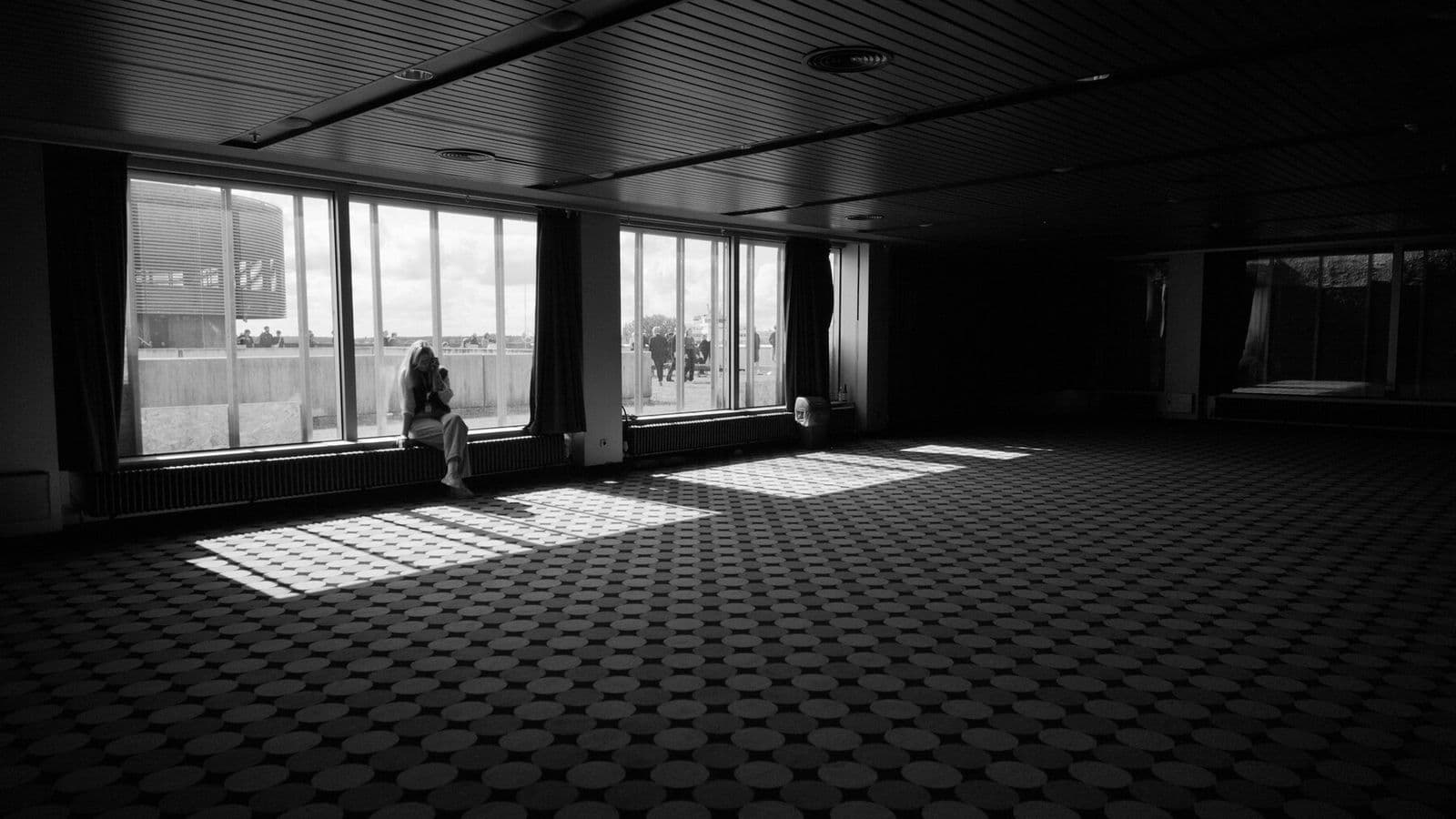 A solitary person sits beside the windows of a large empty room