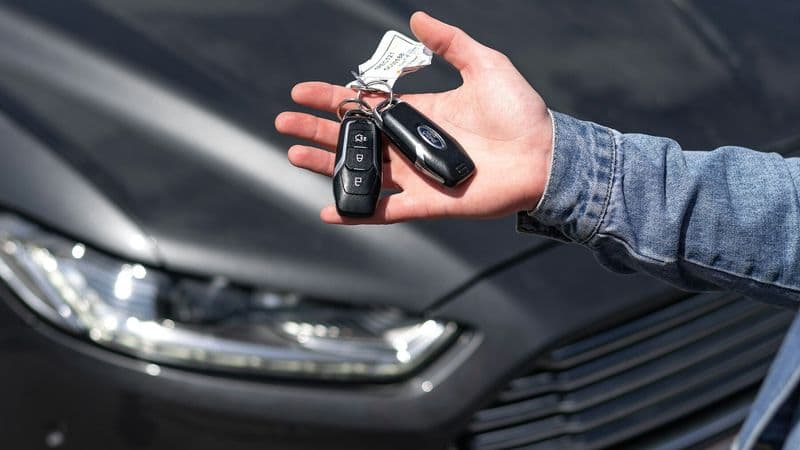 A person holds out a set of car keys beside a parked vehicle